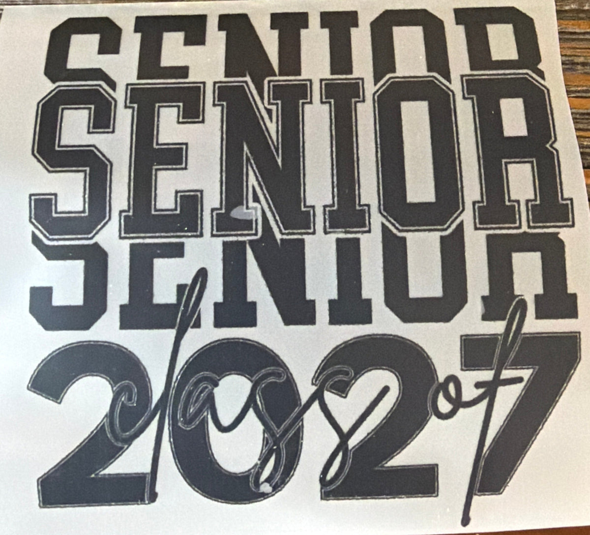 A square white sticker or decal with the text 'SENIOR SENIOR class of 2027' in dark gray lettering, placed on a brown wooden surface.