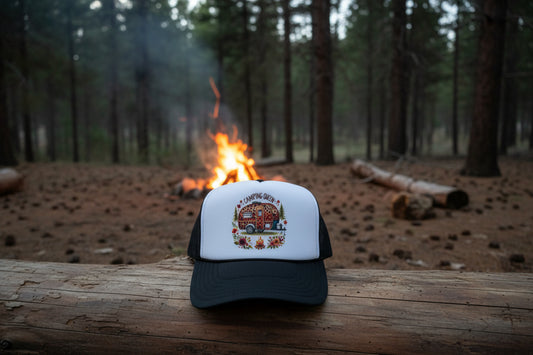A black and white trucker hat with a 'Camping Queen' graphic on the front panel, resting on a rustic wooden surface.