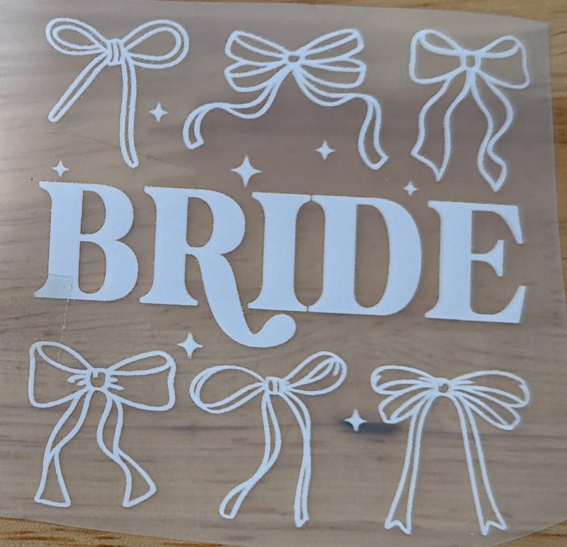 A clear vinyl decal sheet featuring the word 'BRIDE' in white block letters, surrounded by white bow outlines and small star accents, placed on a light wood tabletop.