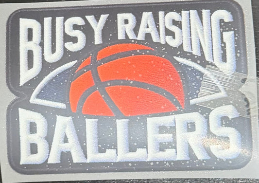 A DTF hat print with the text 'BUSY RAISING BALLERS' and a basketball graphic, placed on a dark wooden surface.