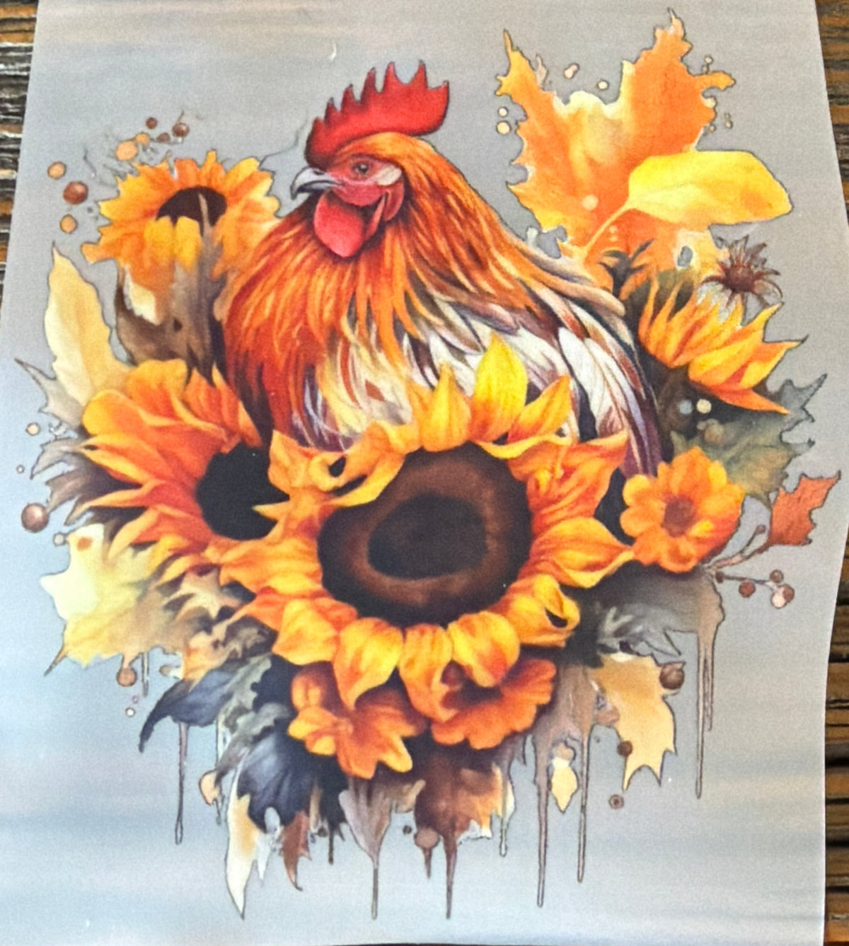 A square art print featuring a watercolor-style rooster surrounded by sunflowers and autumn leaves, placed on a dark wooden surface.