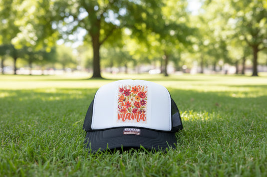 A black and white trucker hat with a colorful floral graphic on the front panel that reads 'mama' in orange script. The hat is displayed on a rustic wooden surface, with another blue and white hat partially visible on the right.