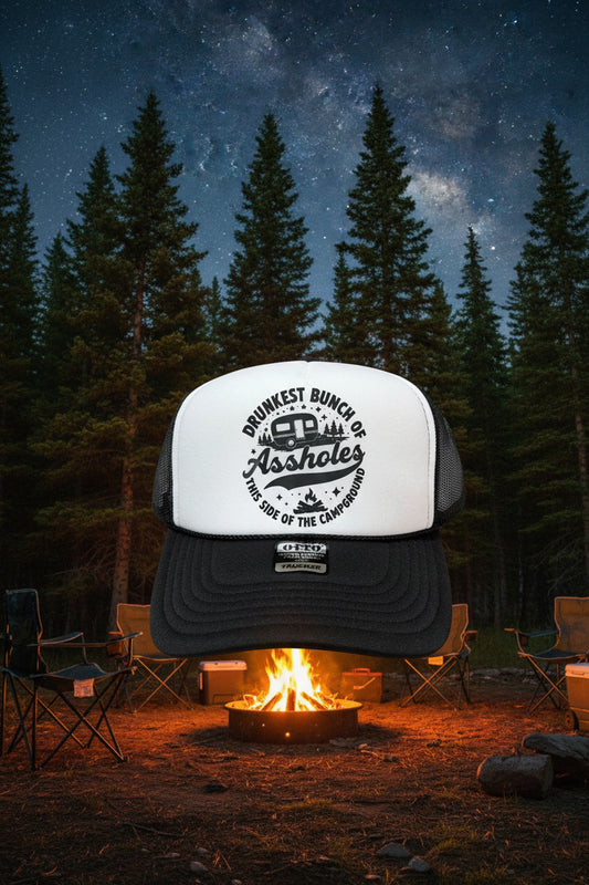 A black and white trucker hat with a white front panel and black mesh back, featuring a black graphic that reads 'DRUNKEST BUNCH OF Assholes THIS SIDE OF THE CAMPGROUND' around a camper illustration. A black brim has a visible 'OTTO' brand sticker.