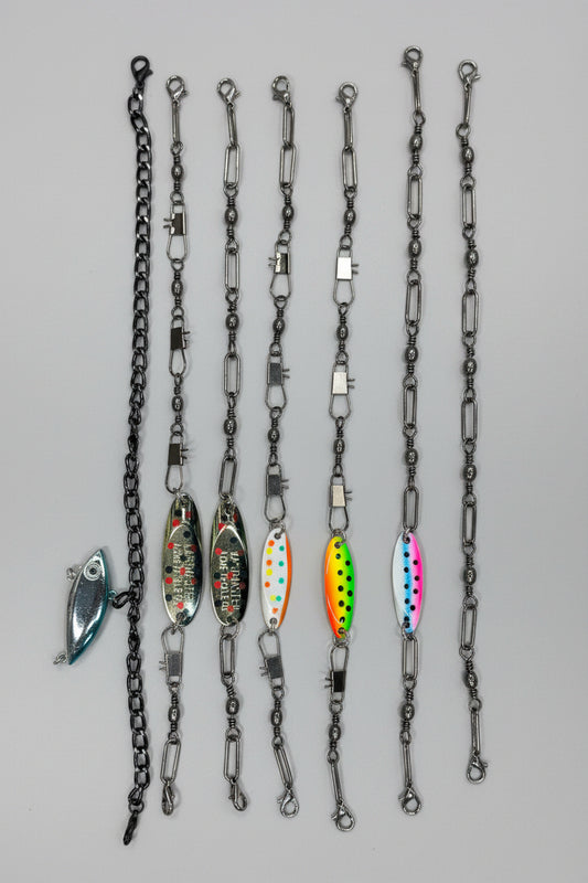 Fishing hat chains with attached dark metal chains are arranged vertically on a light wood surface. The lures include a silver fish-shaped lure, three oval green-and-white patterned lures, and two bright neon lures with pink, orange, yellow, and blue details.