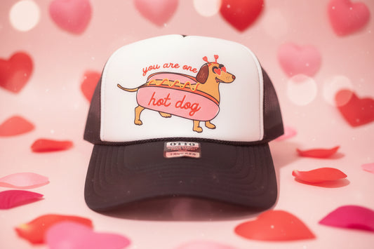 A black and white trucker hat with a cartoon dachshund hot dog graphic on the front panel. The hat has a black brim, a white front panel, and black mesh side/back panels.
