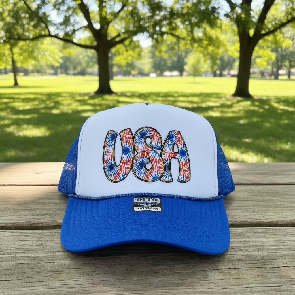 A blue and white trucker hat with a patriotic 'USA' design on the front panel, featuring a red, white, and blue firework pattern. The hat has a royal blue visor and mesh back, with an 'OTTO' brand patch on the brim.