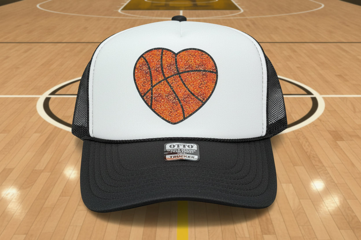 A trucker hat with a white front panel and dark gray mesh back, featuring a glittery orange basketball heart graphic on the front.