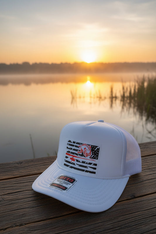 A white trucker hat with a front patch featuring a distressed American flag and a red bass with fishing pole, resting on a dark wooden surface.