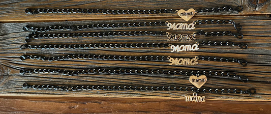 Six black metal link chains with heart-shaped charms engraved with the word 'mama' in gold-tone lettering, arranged on a rustic dark wood background.