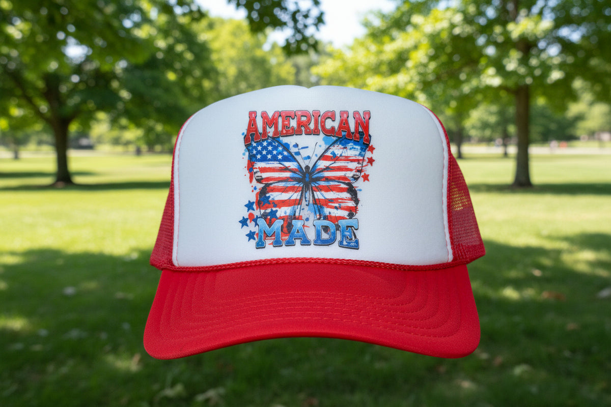A red and white trucker hat with a 'American Made' butterfly graphic featuring the American flag, resting on a wooden table. The hat has a visible OTO logo patch on the brim.