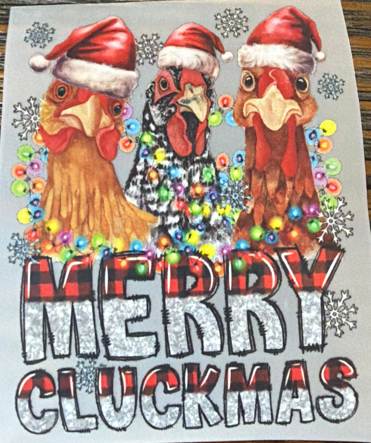 A rectangular printed sticker or decal featuring three cartoon chickens wearing Santa hats, colorful holiday ornaments, snowflakes, and the text 'MERRY CLUCKMAS' in large glitter-style letters, placed on a brown wood surface.