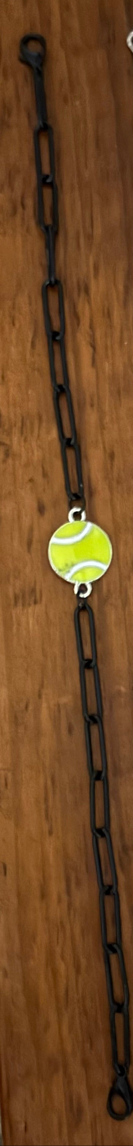 Two tennis-themed bracelets displayed on a wooden surface. One bracelet features a black metal chain with two round lime-green tennis ball charms.