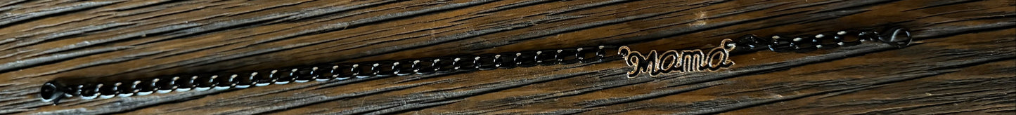 A dark metallic chain bracelet with a small gold-toned script charm spelling MAMA, displayed horizontally on a dark wood background.