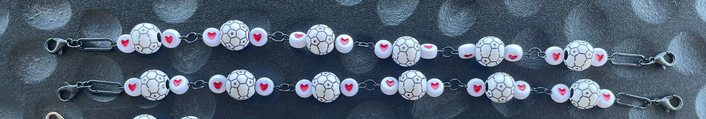 A close-up of a beaded chain bracelet or necklace featuring white oval beads with black crackle patterns, pink heart accents, and silver-toned metal links on a dark textured background.