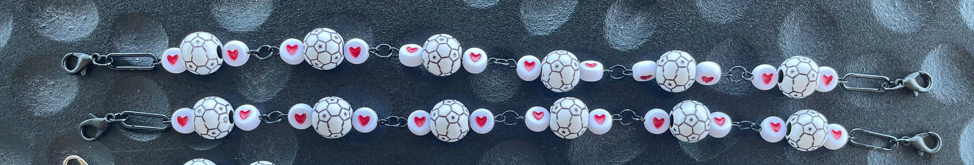 A close-up of a beaded chain bracelet or necklace featuring white oval beads with black crackle patterns, pink heart accents, and silver-toned metal links on a dark textured background.