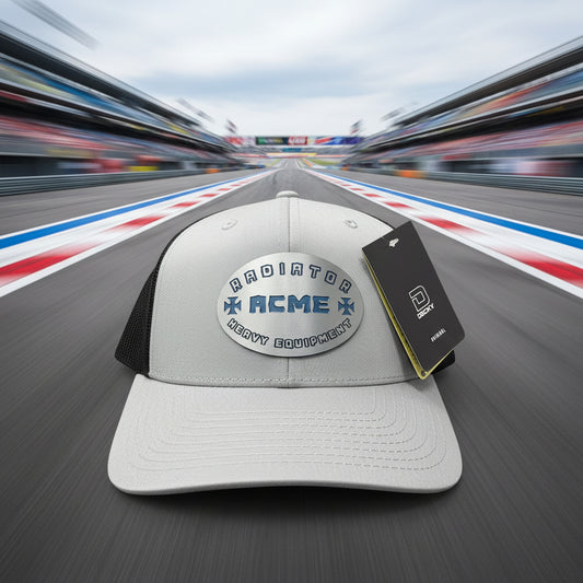 A light gray and black trucker hat with a curved brim, featuring a metallic silver oval patch with blue text that reads 'RADIATOR ACME HEAVY EQUIPMENT' and a black hangtag and sticker on the brim.