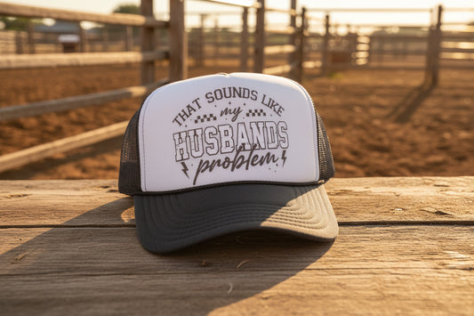 A dark gray and white trucker hat with a printed graphic on the front panel that reads 'THAT SOUNDS LIKE my HUSBANDS problem' in gray lettering. The hat features a white front panel, dark gray mesh back, and a dark gray brim.