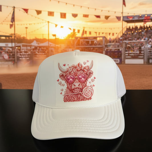A white trucker hat with a pink and red embroidered Scottish highland cow graphic on the front panel. The hat features a white foam front panel, white curved brim, and white mesh back panels.