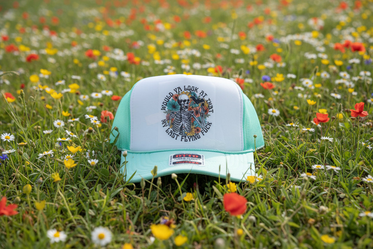 A mint green and white trucker hat with a graphic print of a skeleton and flowers on the front panel.