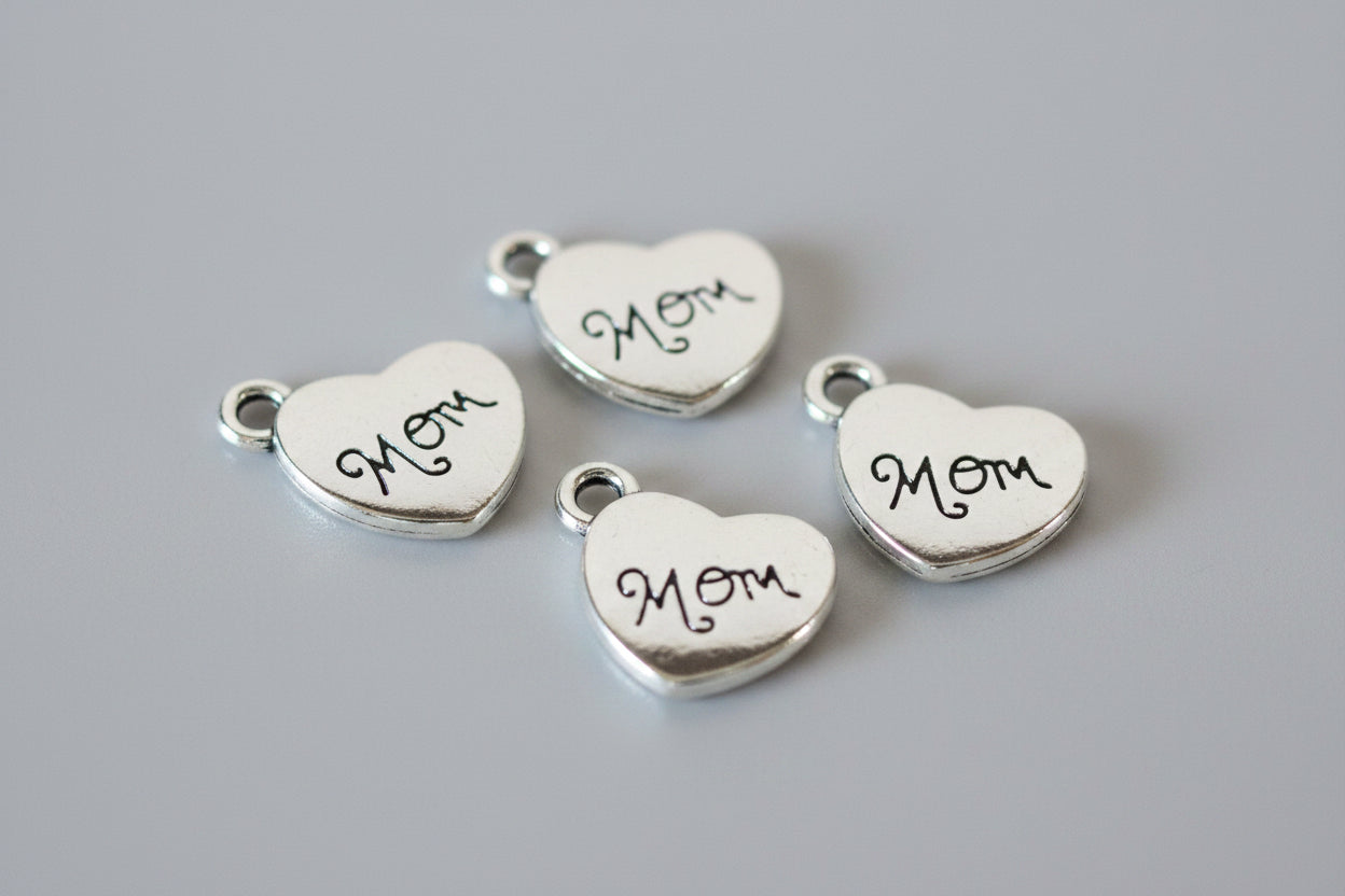 Four silver heart-shaped charms engraved with 'Mom' and topped with loops, arranged on a dark wood background.