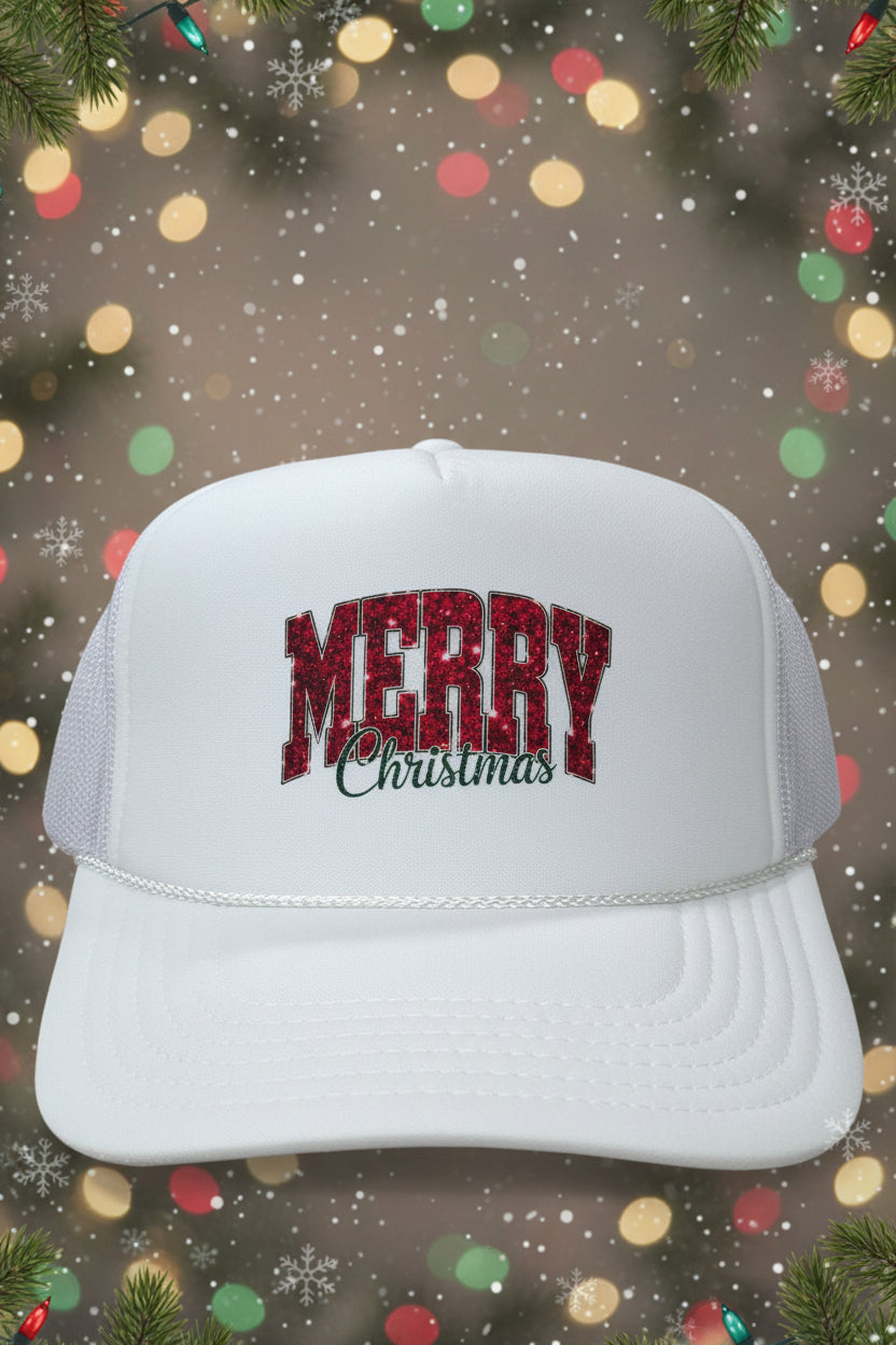 A white trucker hat with 'MERRY Christmas' on the front, resting on a wooden table. The hat features a red and black glittery 'MERRY' text with 'Christmas' in black cursive underneath.