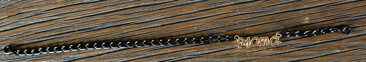 A black metal chain bracelet with a small gold-toned 'MAMA' script charm, displayed horizontally on a dark wood background.