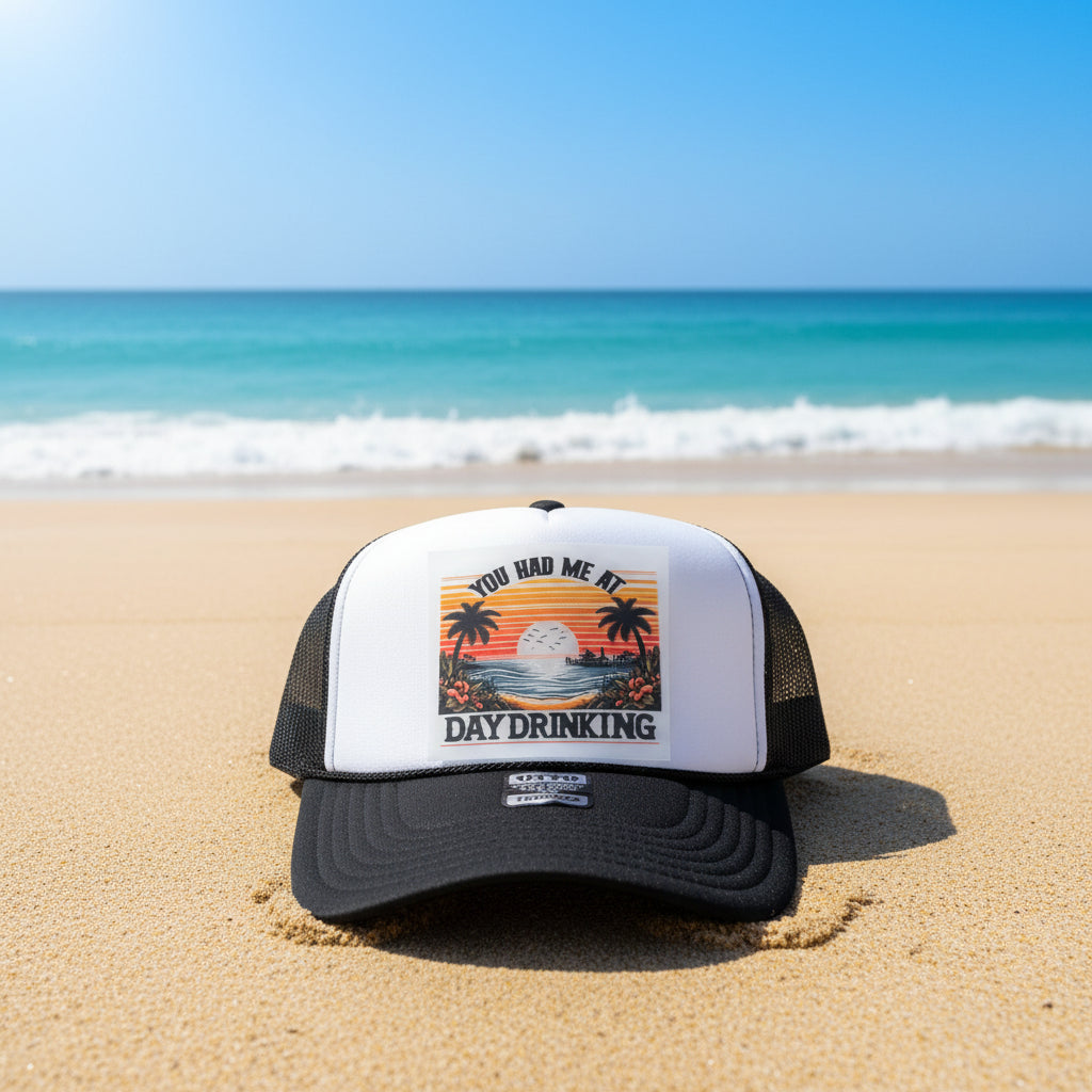 A black and white trucker hat with a retro sunset graphic on the front panel that reads 'YOU HAD ME AT DAY DRINKING'. The hat has a black brim, black mesh sides, and a white front panel.