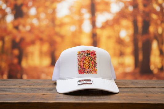 A white foam trucker hat with a colorful floral pumpkin patch graphic on the front, resting on a dark wooden surface. The hat features a visible 'OTTO' brand patch on the brim.
