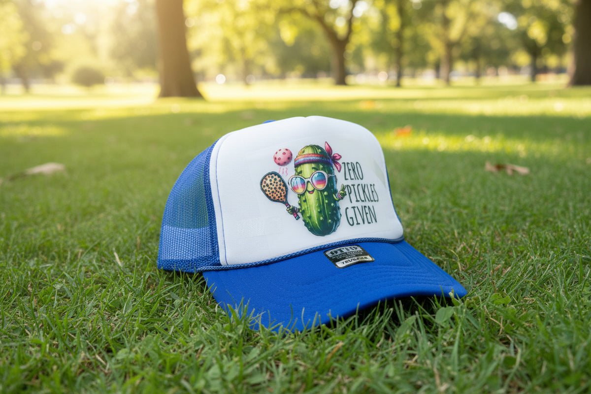 A blue and white trucker hat with a graphic of a pickle character and the text 'ZERO PICKLES GIVEN' displayed on a wooden table, with other hats in the background.