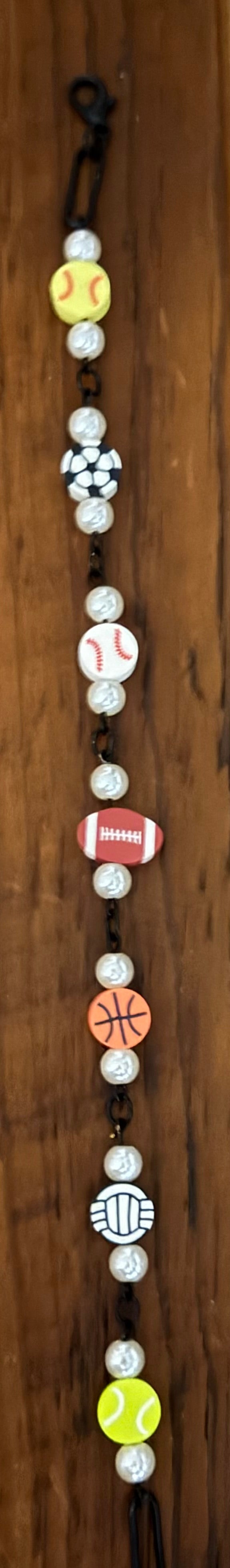 A beaded necklace featuring sports-themed beads including a yellow baseball, a black-and-white checkered pattern, white baseballs, a red-and-white football, an orange basketball, a black-and-white checkerboard pattern, and a yellow tennis ball, displayed on a dark wood background.