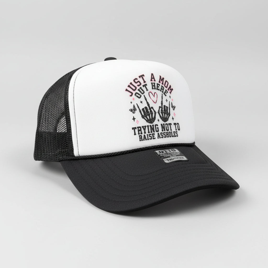A black and white trucker hat with a graphic on the front panel that reads 'JUST A MOM OUT HERE TRYING NOT TO RAISE ASSHOLES' with two black hand gestures and a small pink heart. A metal GTT sticker is visible on the brim.