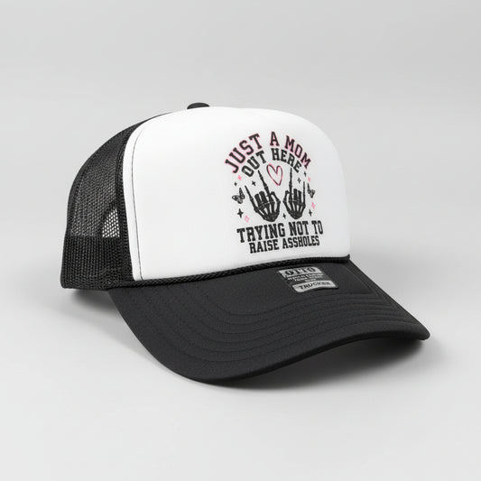 A black and white trucker hat with a graphic on the front panel that reads 'JUST A MOM OUT HERE TRYING NOT TO RAISE ASSHOLES' with two black hand gestures and a small pink heart. A metal GTT sticker is visible on the brim.