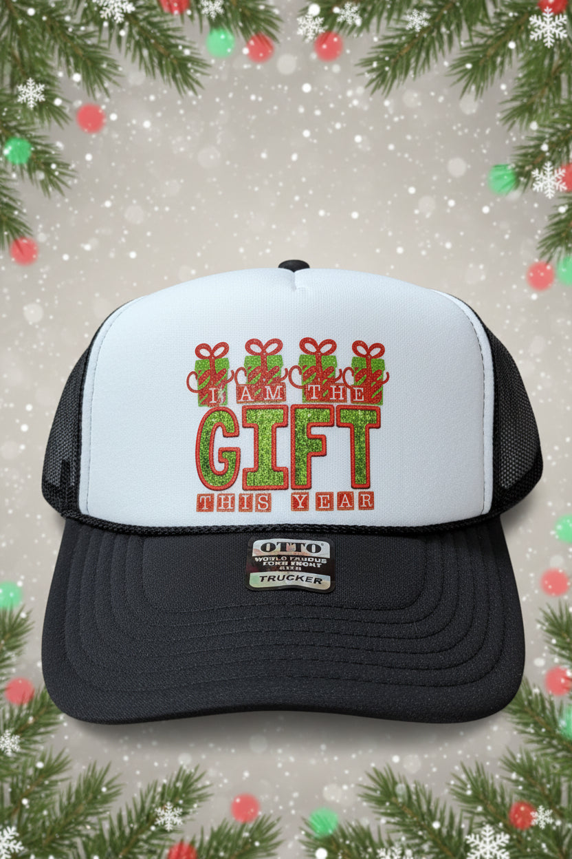 A black and white trucker hat with a custom holiday graphic on the front panel that reads 'I AM THE GIFT THIS YEAR' in red and green glittery lettering. The hat has a black brim and black mesh back, with an Otto brand sticker on the brim.