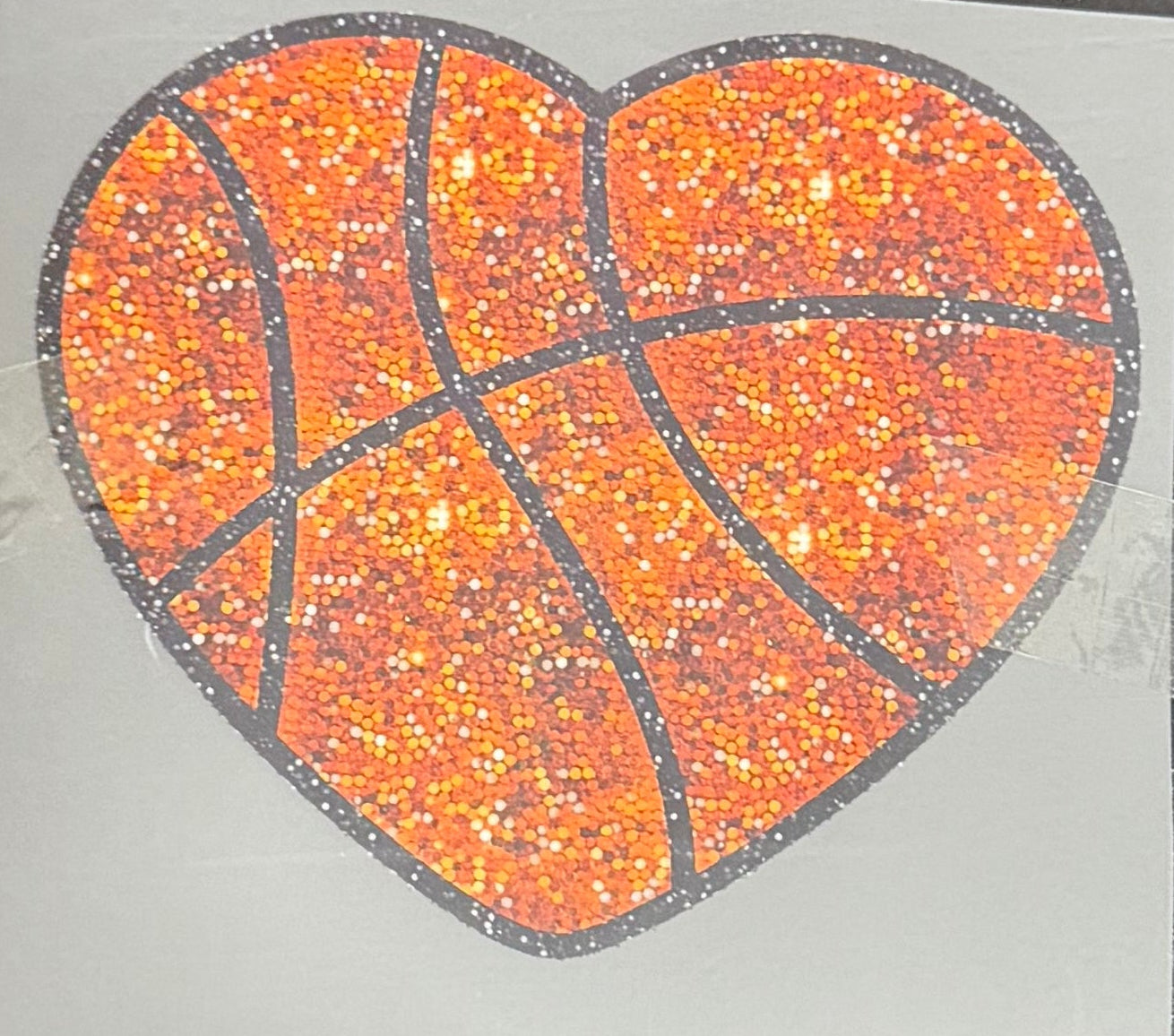 A square white sticker or card featuring a glittery orange basketball heart design, taped down on a dark wooden table.