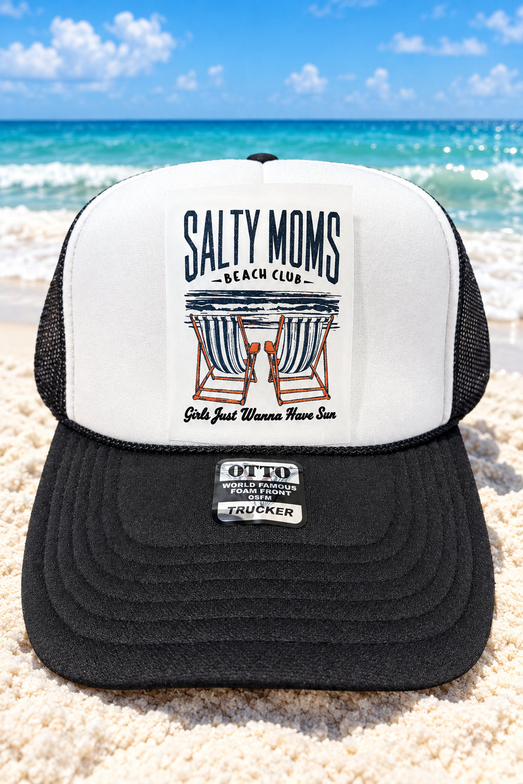 A black and white trucker hat with a white front panel and black brim, featuring a 'Salty Moms Beach Club' graphic and an OTK brand patch on the brim, resting on a wooden surface.
