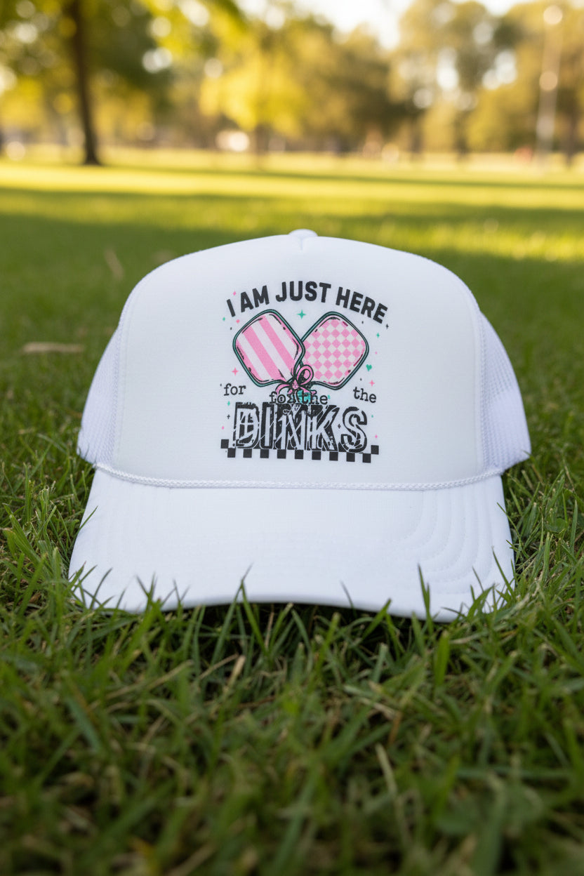 A white trucker hat with a graphic on the front that reads 'I AM JUST HERE for the DINKS' with two pink patterned paddles. A visible patch on the brim reads 'OTTO World Famous Foam Front OSFM Trucker'.