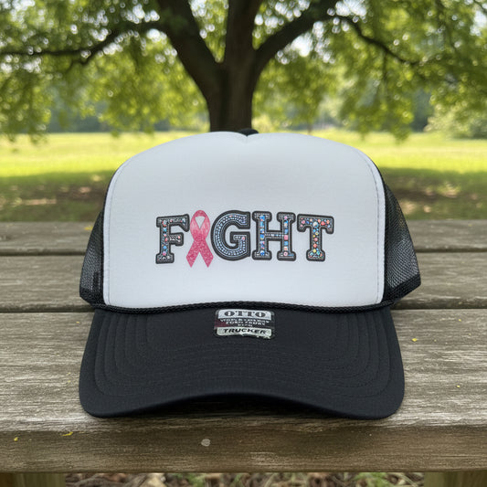 A white and black trucker hat with 'FIGHT' lettering on the front, where the 'I' is replaced by a pink ribbon. The hat has a white front panel, black mesh back, and a black brim with a visible 'OTTO' brand tag.