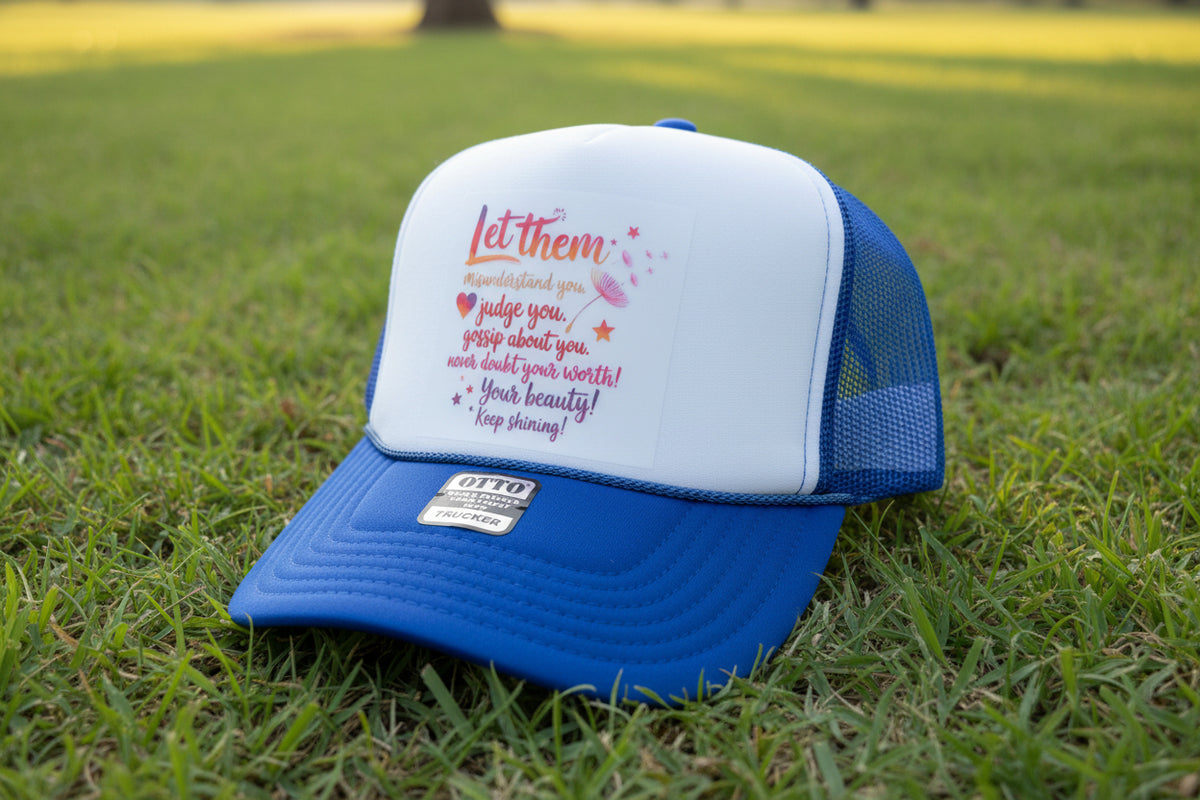 A blue and white trucker hat with a printed motivational quote on the front panel. The hat features a royal blue brim and mesh back, with a white front panel printed with the text 'Let them misunderstand you judge you gossip about you never doubt your worth! Your beauty! Keep shining!' in pink, orange, and red lettering.