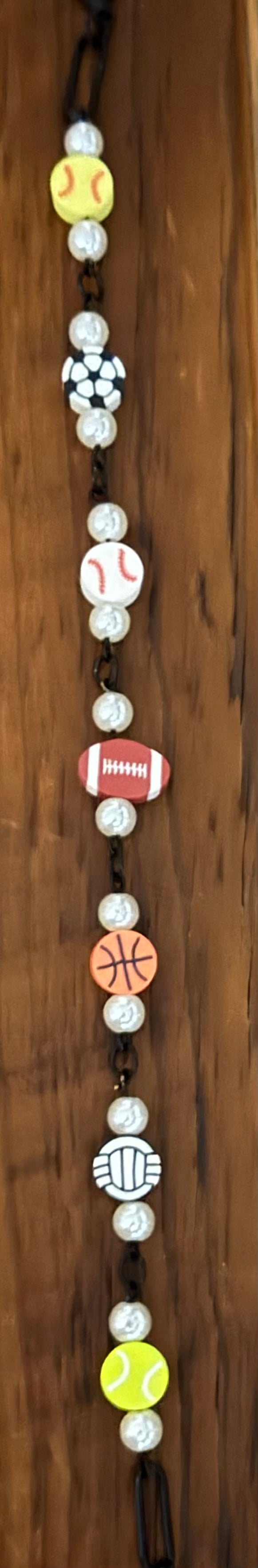 A beaded necklace featuring sports-themed beads including a yellow softball, white baseball, black-and-white soccer ball, red football, orange basketball, white volleyball, and yellow tennis ball, strung on a dark cord against a wooden background.
