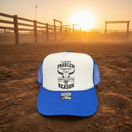 A blue and white trucker hat with a black graphic print on the front, placed on a dirt ground with a ranch fence and sunset in the background.