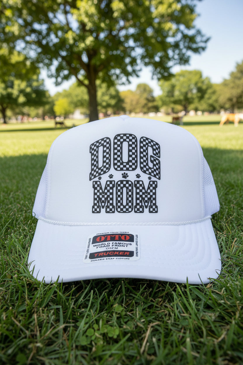 A white trucker hat with a 'DOG MOM' graphic on the front panel, resting on a wooden surface.