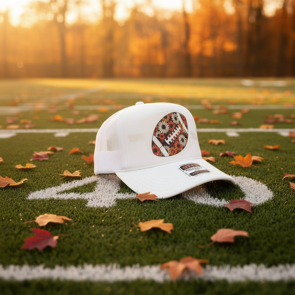 A white trucker hat with a floral football patch on the front, resting on a black table against a dark wood background.
