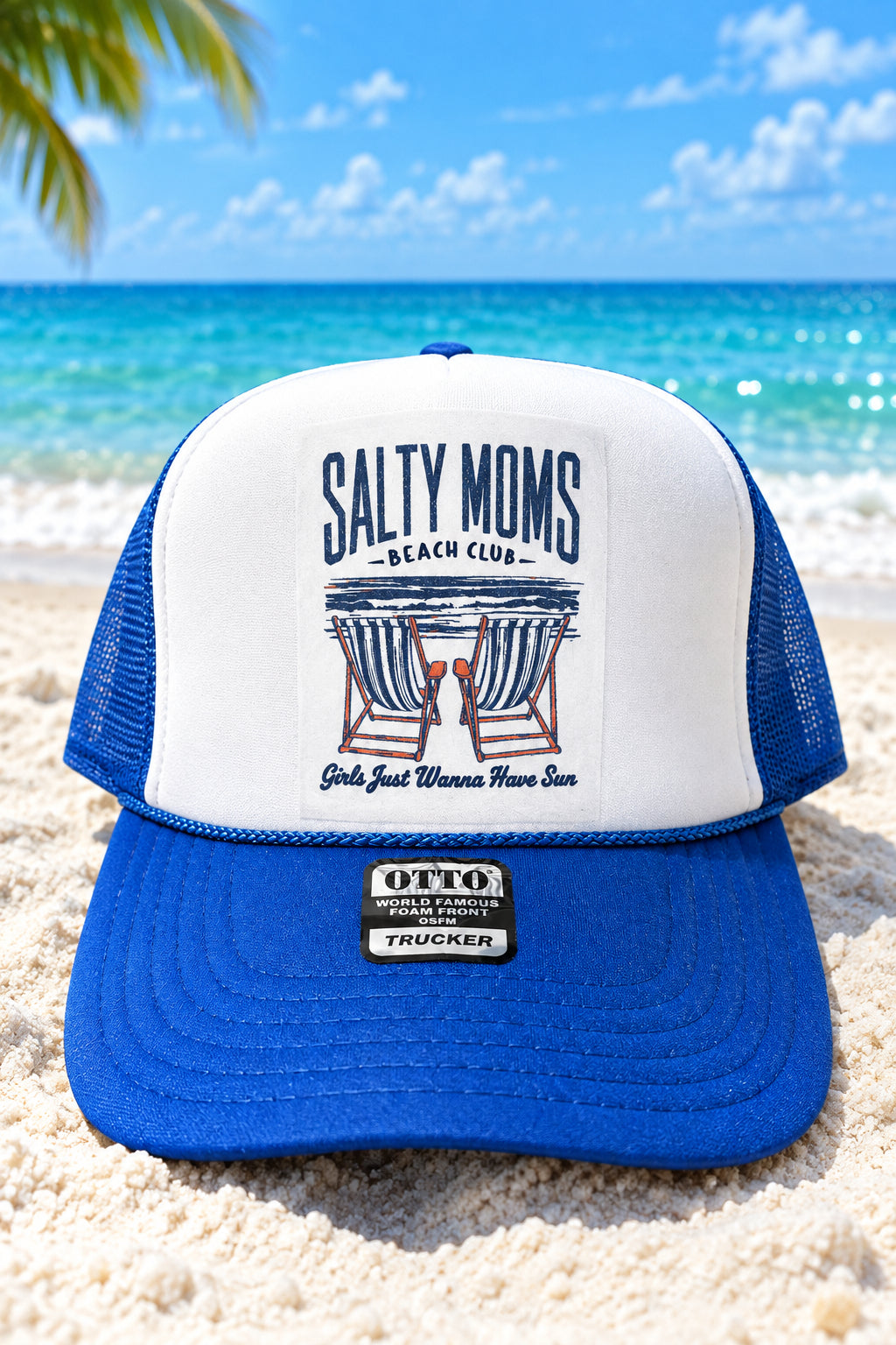 A blue and white trucker hat with a white front panel and blue mesh sides, featuring a 'Salty Moms Beach Club' graphic on the front and an 'Otto' trucker patch on the brim, displayed on a rustic wooden surface.