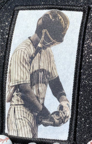A black glittery baseball cap with a baseball-themed beaded headband, held up by a hand against a blurred baseball field background.