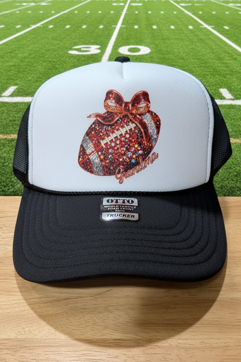 A black and white trucker hat with a custom glittery football graphic on the front panel. The graphic features a red glitter football with a red glitter bow and the word 'Grandma' in red glitter script.