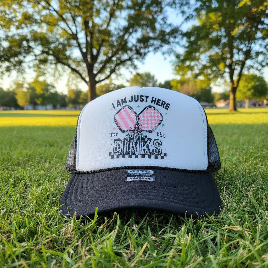 A black and white trucker hat with a graphic on the front panel that reads 'I AM JUST HERE. for the dinks' with two pink patterned bowties.