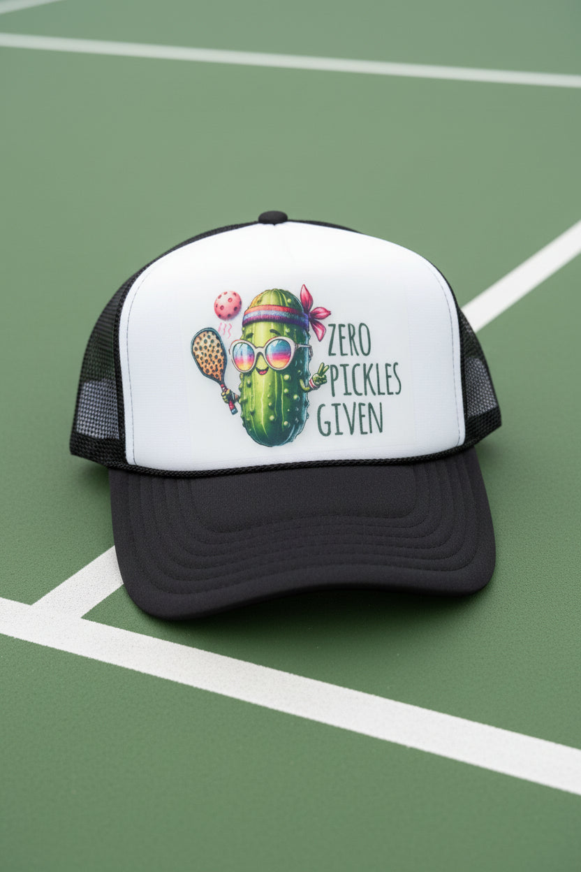 A black and white trucker hat with a graphic of a pickle character wearing sunglasses and a headband, next to the text 'ZERO PICKLES GIVEN'. The hat is resting on a wooden surface with a blurred background.