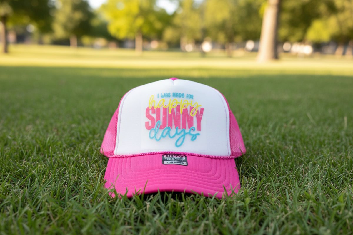 A pink and white trucker hat with a white front panel and bright pink brim, featuring the text 'I WAS MADE FOR happy SUNNY days' in colorful script. The hat has a visible Otto Trucker brand patch on the brim and is photographed against a dark wood background.