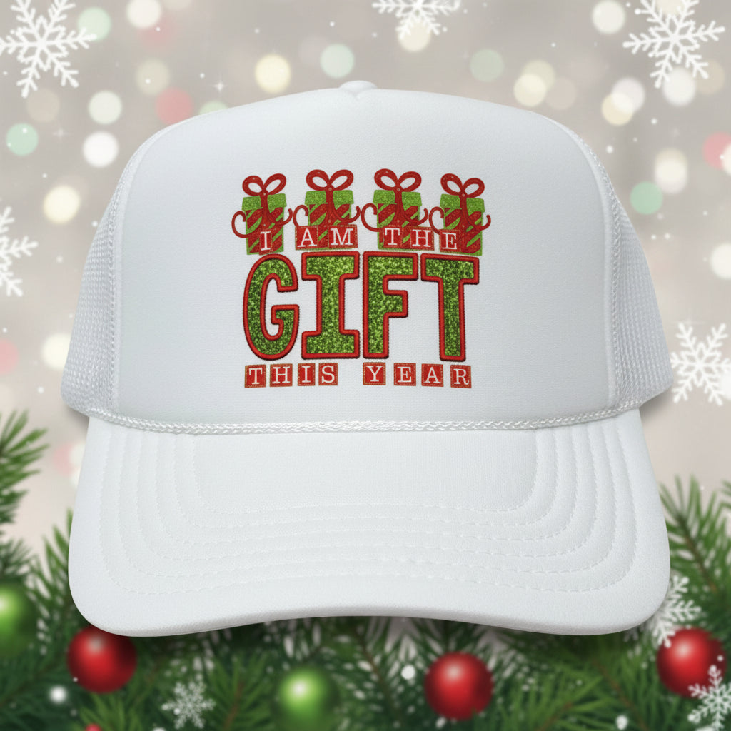 A white trucker hat with a festive holiday graphic on the front that reads 'I AM THE GIFT THIS YEAR' in red and green glitter-style lettering. The hat has a white foam front and white mesh back, and a sticker on the brim reads 'OTTO'.