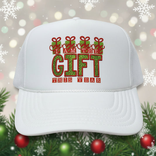 A white trucker hat with a festive holiday graphic on the front that reads 'I AM THE GIFT THIS YEAR' in red and green glitter-style lettering. The hat has a white foam front and white mesh back, and a sticker on the brim reads 'OTTO'.